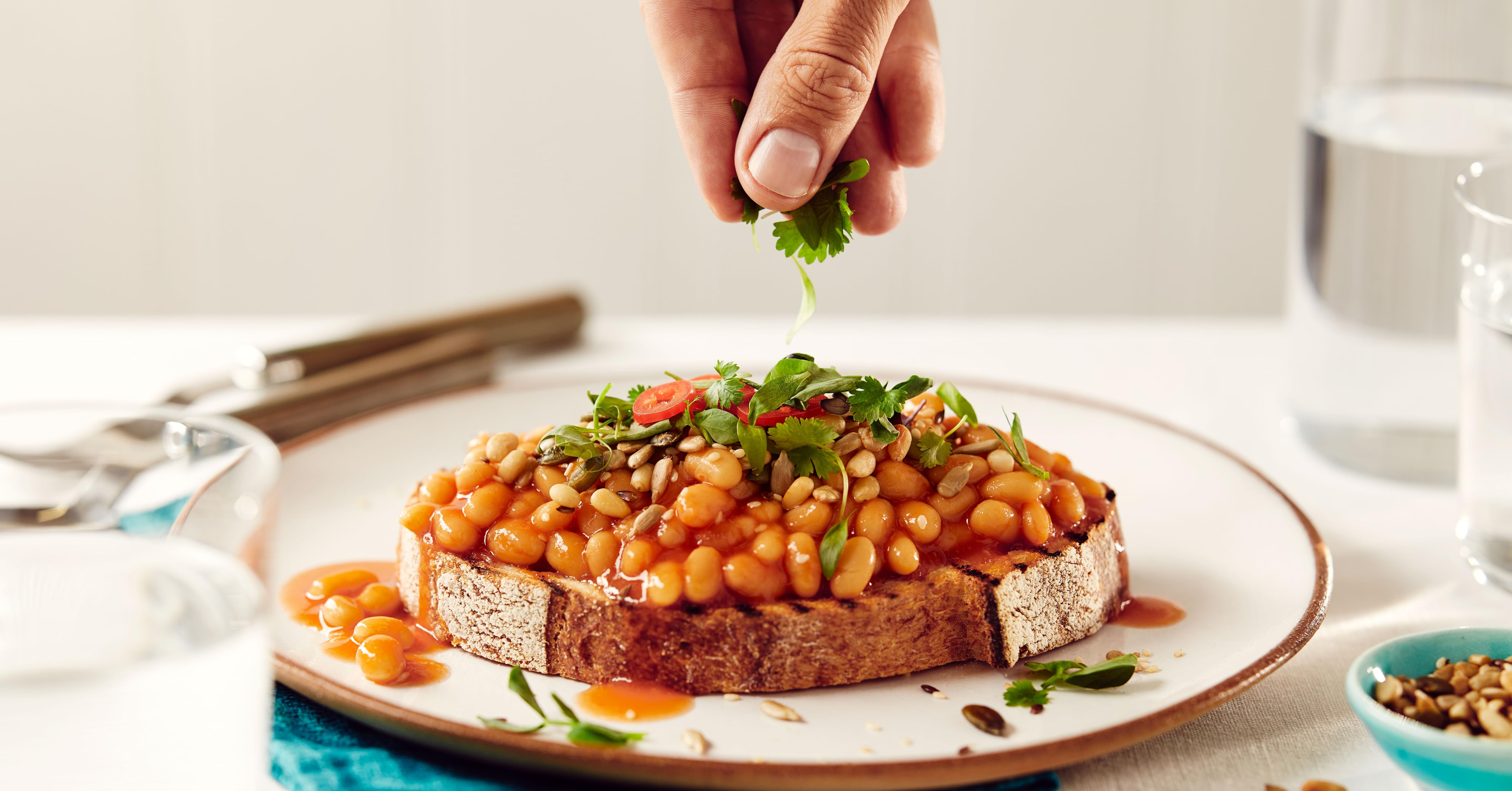 Plate with toast topped with baked beans, garnished with herbs. Hand sprinkling additional herbs over the dish.