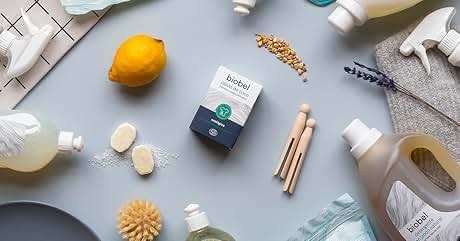 Surtido de productos de limpieza y cuidado personal sobre superficie gris. Incluye limón, pinzas de madera, cepillos, botellas de aerosol y artículos