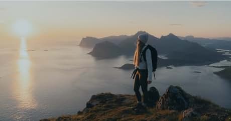 El texto dice «Imagen 1". La persona con la mochila está parada en la cima de la montaña con vistas al paisaje costero con islas al atardecer. Silueta de la figura contra el cielo anaranjado y el agua reflectante.