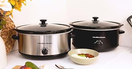 Two slow cookers on a kitchen counter. One is stainless steel, the other black. A bowl of soup and fresh vegetables are visible nearby.