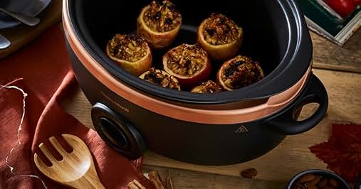 Slow cooker with copper-colored band containing baked apples stuffed with nuts. Wooden utensils, red napkin, cookbook, whole apples, and bowl of almonds visible in background.