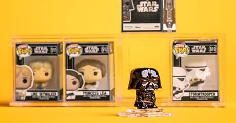 de Star Wars se muestran sobre fondo amarillo. Figuras de Funko Pop en cajas y una pequeña figura de Darth Vader en la parte delantera. Póster de Star Wars visible