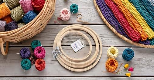 Des cerceaux à broder en bois de différentes tailles entourés d'écheveaux et de boules de laine colorés. Un panier en osier contient de la laine supplémentaire. Fournitures d'artisanat disposées sur une surface en bois clair