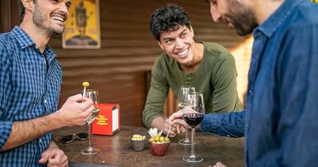 Tres personas se reunieron alrededor de una mesa con copas de vino, bocadillos y una caja roja, disfrutando de una reunión social en el interior.