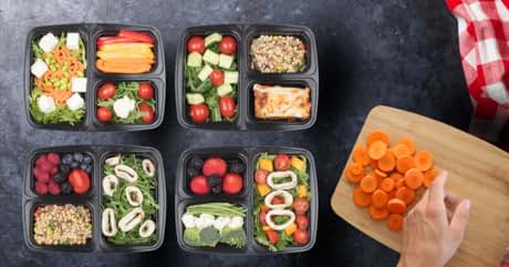 Eight black meal prep containers filled with various colorful foods. Vegetables, fruits, and grains visible in separate compartments.