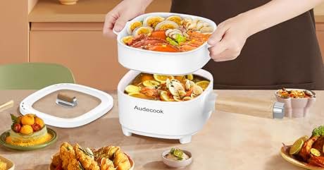 White electric food steamer with two stacked compartments. Top layer contains seafood and vegetables, bottom layer has pasta dish. Surrounding plates show various cooked foods.