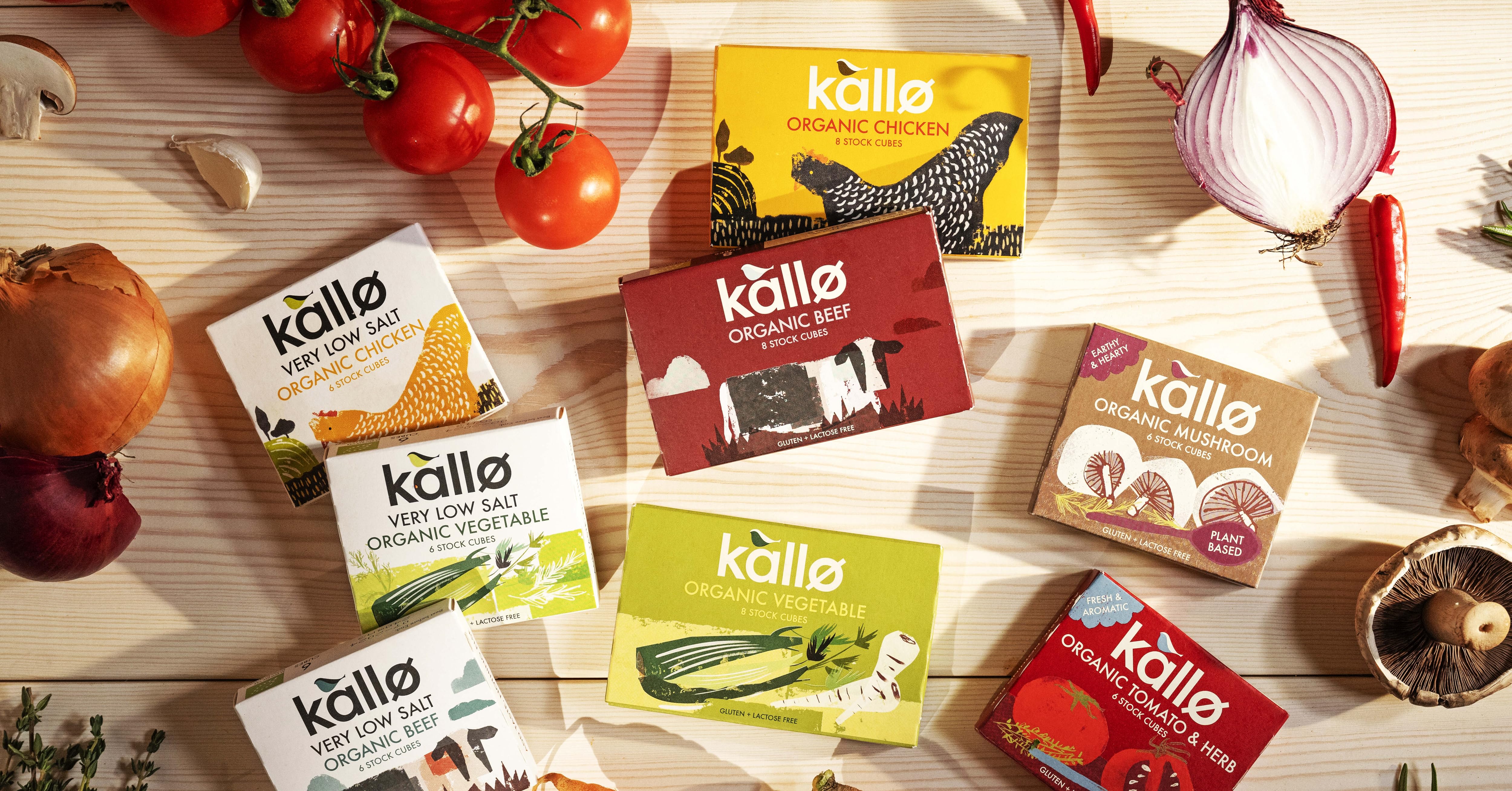 Various Kallo brand food product boxes displayed on a wooden surface with fresh vegetables like tomatoes, carrots, and garlic.