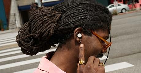 Primer plano de una persona con rastas que lleva pendientes dorados y gafas de sol, sujetándose un dispositivo a la oreja al aire libre
