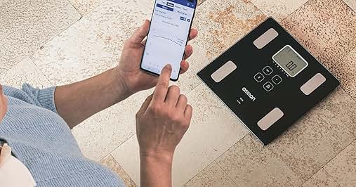 Manos que sostienen el teléfono inteligente junto a una báscula corporal digital sobre un suelo de baldosas. La báscula tiene una pantalla digital y varios sensores visibles en su superficie