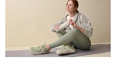 Frau sitzt auf einer Gymnastikmatte und trägt hellgrüne Leggings, grauen Hoodie und mintgrüne Turnschuhe. Sie hält eine helle Schaumstoffrolle gegen
