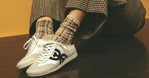 Text reads "SNEAKERS". White sneakers with black logo, worn with plaid pants and matching socks. Person sitting on wooden surface against orange background.