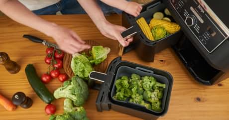 Kitchen scene with hands using an air fryer. Various vegetables on wooden surface. Air fryer contains broccoli. Other kitchen appliance visible.