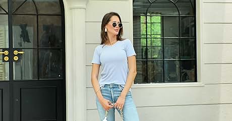Mujer vestida con camiseta azul claro, jeans y gafas de sol parada frente a un edificio blanco con grandes ventanales y puerta negra.