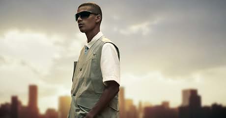 con chaleco sin mangas beige sobre camisa blanca con el horizonte urbano como telón de fondo. Gafas de sol oscuras y pelo corto completan el look