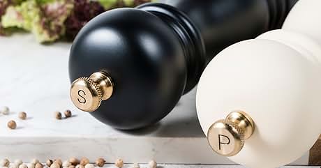 Large black and white pepper mills on a surface with scattered peppercorns. Elegant, cylindrical design with metal accents.