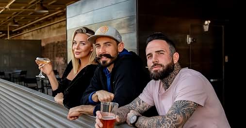 Three people sitting at outdoor bar, holding drinks. One person gesturing while others listen.
