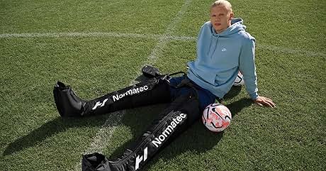 Mangas de compresión negras para piernas con la etiqueta «NormaTec» que lleva una persona sentada en el césped con un balón de fútbol, demostrando su equipo de recuperación deportiva