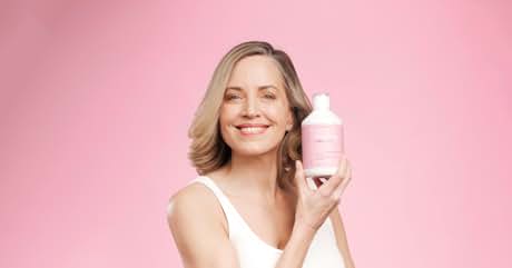 Mujer con blusa blanca que sostiene una botella rosa de producto para el cabello o el cuidado de la piel sobre un fondo rosa. Parece estar promocionando el producto
