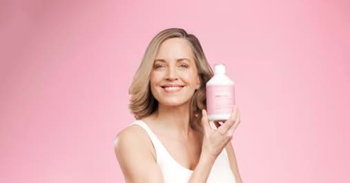 Mujer con blusa blanca que sostiene una botella rosa de producto para el cabello o el cuidado de la piel sobre un fondo rosa. Parece estar promocionando el producto