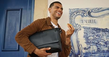 El texto dice «MONEHIQUE». Hombre que sostiene una funda gris para portátil, sonriendo. Llevaba una chaqueta marrón. Puerta azul y azulejos decorativos con paisaje urbano visible en el fondo