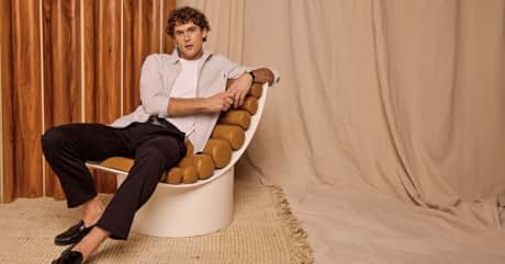 Persona con camisa blanca y pantalones oscuros sentada en una silla de diseño único con asiento curvo y acolchado sobre un fondo claro