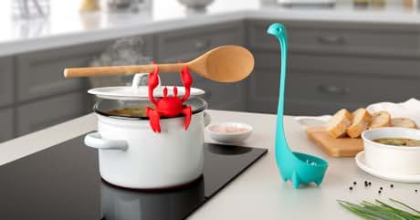 Text reads "Image 1". Kitchen scene with whimsical utensils: red crab-shaped steam releaser on white pot, turquoise dinosaur-shaped ladle nearby. Wooden spoon, bread slices, and seasonings visible on countertop.
