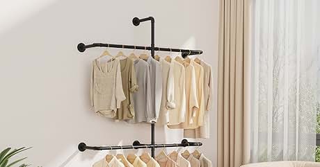 Perchero doble montado en la pared con tubos de metal negro. En ambos niveles cuelgan prendas de colores neutros. Sala de estar con sofá, cortinas, piso de madera y accesorios a la vista, como bolsos y zapatos