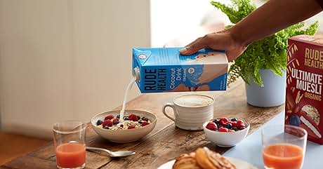 Hand pouring a blue carton of plant-based milk over a breakfast setting with berries in bowls, pastries on a plate, orange juice glasses, and a cereal box on a wooden table.