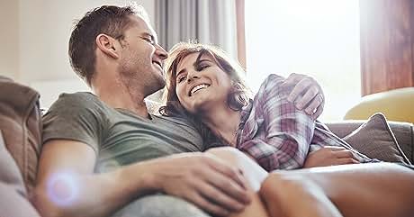 Pareja relajándose en el sofá. Hombre con camisa verde, mujer con camisa a cuadros. La luz del sol entra por la ventana. Postura íntima y cómoda.