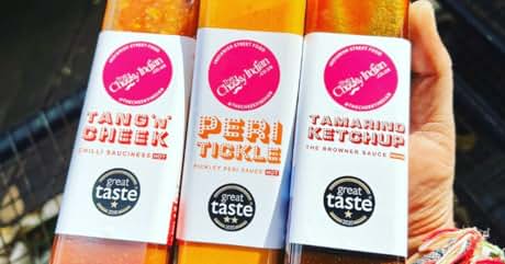 Three glass bottles of colorful sauce: Tangy Cheek (orange), Peri Tickle (yellow), and Tamarind Ketchup (red-orange). Each bottle features a pink circular label and a 'Great Taste' award seal.