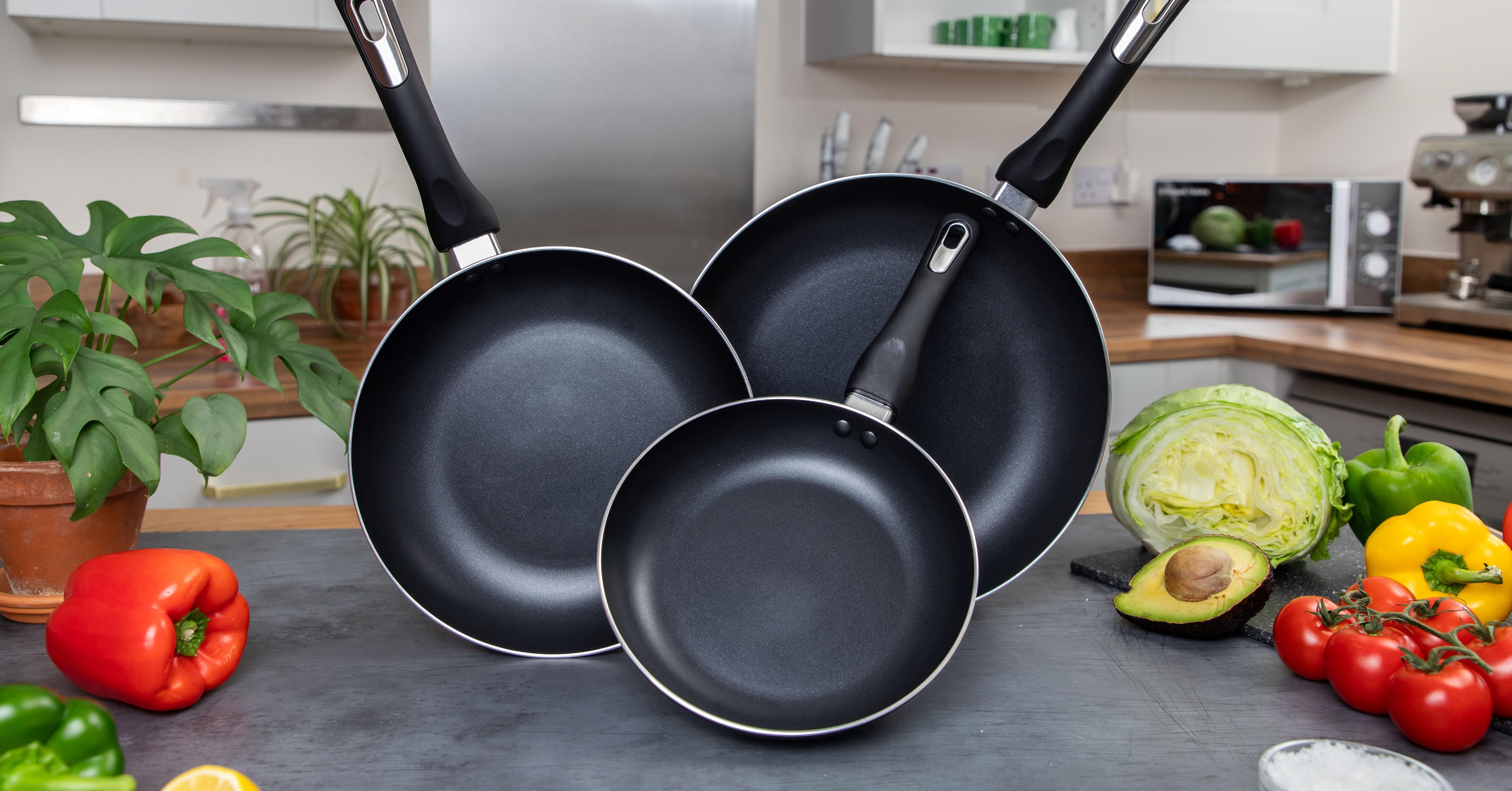 Set of three non-stick frying pans in graduating sizes with black handles, displayed on a counter with fresh vegetables in background.
