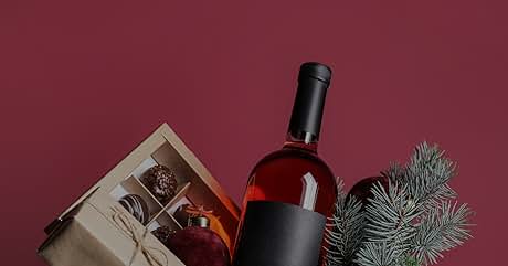 con etiqueta oscura junto a una caja de regalo que contiene chocolates y una rama de pino de hoja perenne sobre fondo burdeos