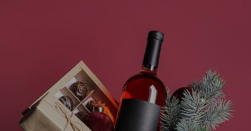 con etiqueta oscura junto a una caja de regalo que contiene chocolates y una rama de pino de hoja perenne sobre fondo burdeos