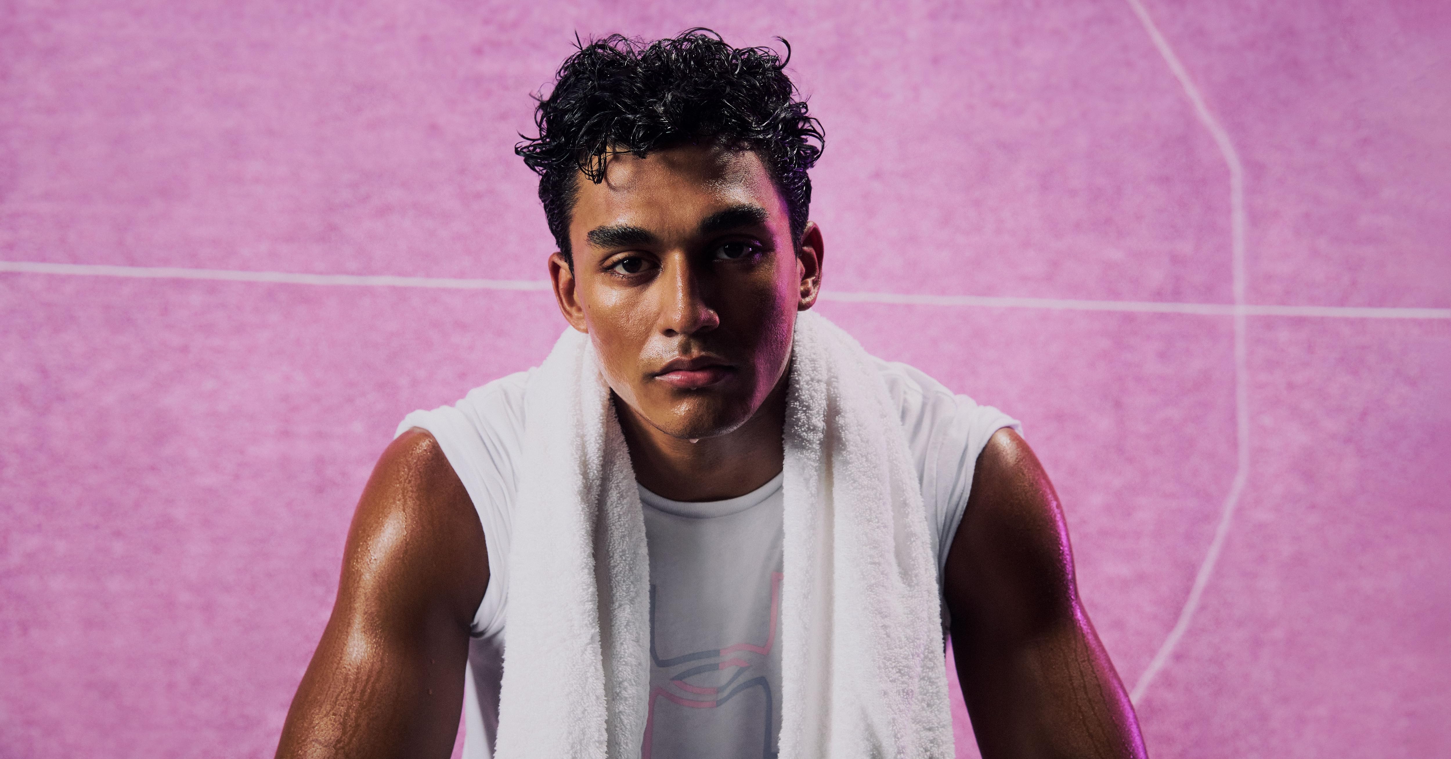 Hombre atlético vestido con camisa blanca sin mangas con toalla alrededor del cuello sobre fondo rosa. El logo de Nike es visible en la camiseta. Brazos musculosos y expresión intensa
