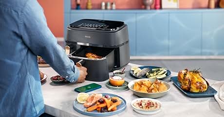 Luftfritteuse auf einer Küchentheke mit verschiedenen gekochten Lebensmitteln, darunter Pommes, Hühnchen und Gemüse