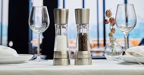 Table setting with two stainless steel and glass salt and pepper grinders in center. Empty wine glasses, white plates, and cutlery visible. Large windows in background show outdoor view.