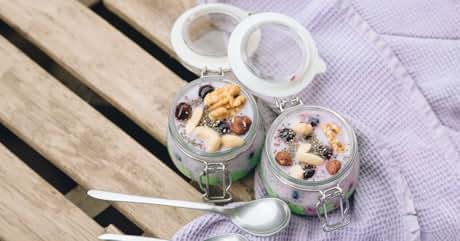 Two glass jars with clip-top lids containing yogurt or pudding topped with berries and nuts. Spoons and a purple cloth are visible on a wooden surface.