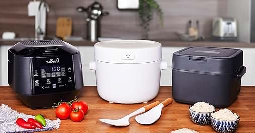 Three kitchen appliances on wooden counter: black pressure cooker with digital display, white rice cooker, and dark gray slow cooker. Ingredients and utensils surrounding them.