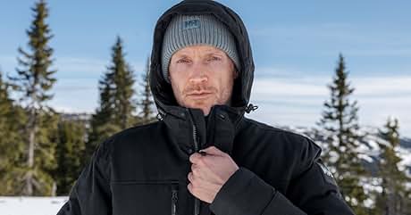 Hombre vestido con chaqueta de invierno negra con capucha y gorro de punto gris en un paisaje montañoso cubierto de nieve con árboles de hoja perenne y un cielo azul claro