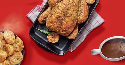 Roasted whole chicken on black tray with potatoes and herbs. Checkered napkin underneath. Side of roasted potato chunks and steaming mug of beverage on red background.