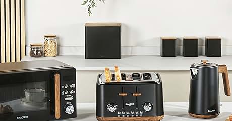 Küchentheke mit schwarzen Geräten und Holzakzenten: Mikrowelle, 4-Scheiben-Toaster und Wasserkocher. In den Regalen darüber befinden sich Küchenutensilien, Bücher und Pflanzen. Kaffeetasse und Gebäck auf der Theke.