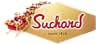de Suchard en forma hexagonal dorada con texto rojo, con ilustración decorativa de trineo. Se muestra la fecha de fundación «desde 1826»
