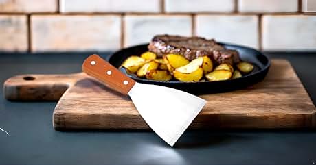 Professional kitchen setup with white triangular spatula featuring wooden handle, placed on wooden cutting board next to cast iron pan with cooked food.