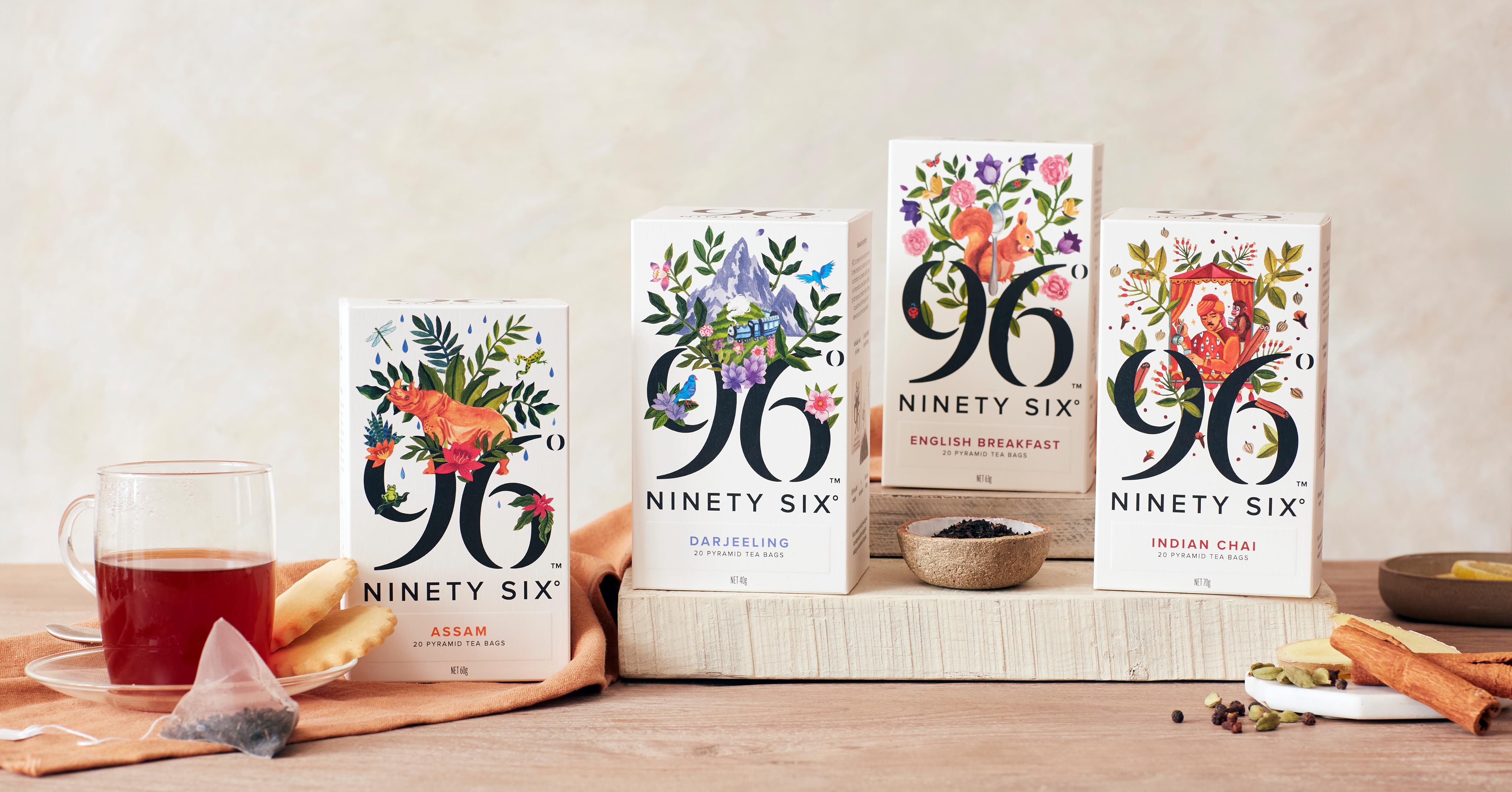 Four boxes of Ninety Six tea displayed on a wooden surface, featuring colorful floral designs. Accompanying the boxes are a cup of tea, a tea bag, and various spices.