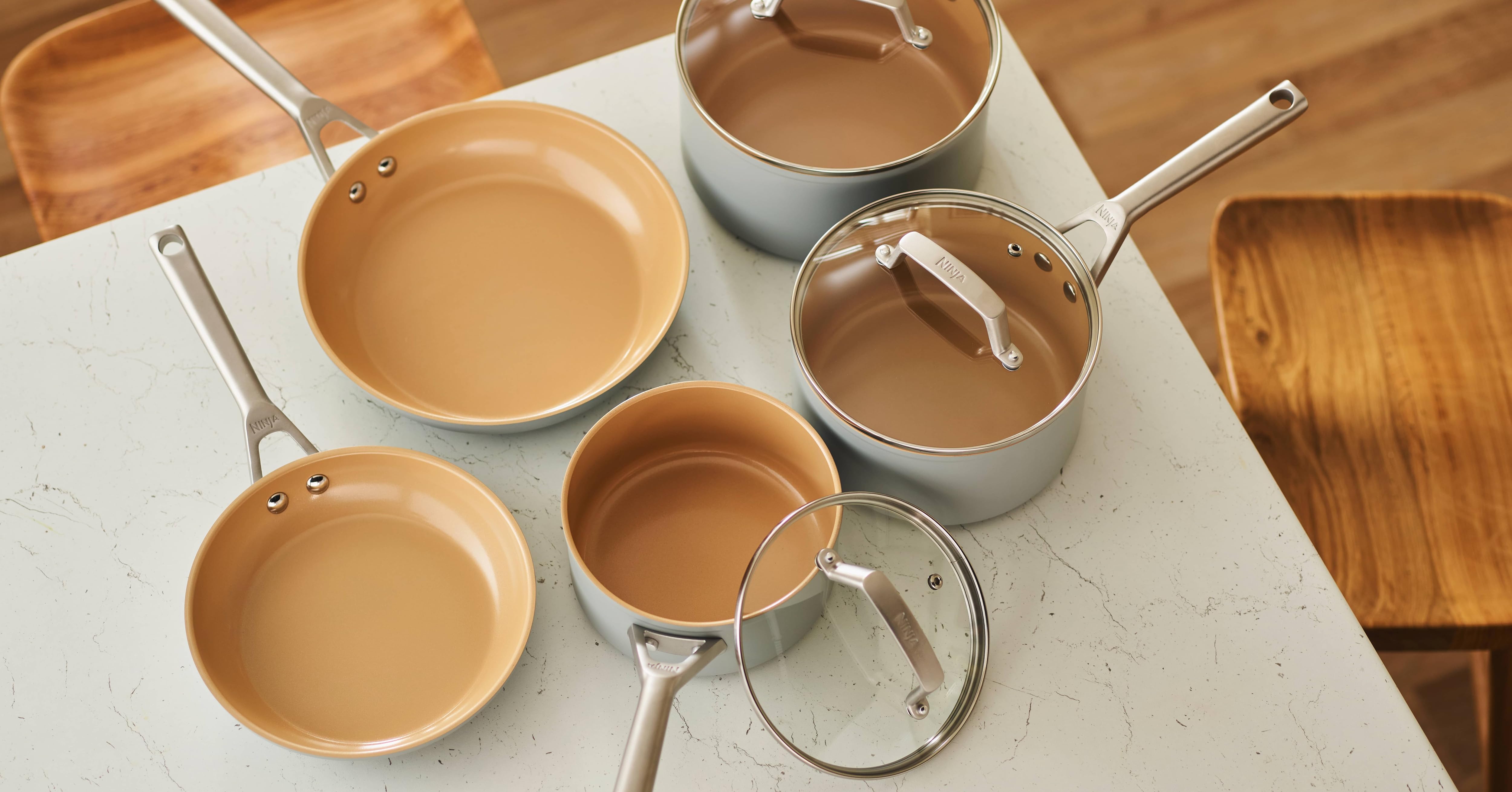 Set of ceramic-coated cookware on white surface. Collection includes frying pans and saucepans in various sizes with beige interior and light gray exterior. Stainless steel handles and glass lid visible.