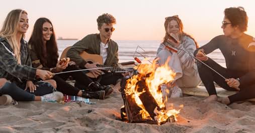 Beach bonfire scene at sunset with group gathered around campfire on sandy shore.