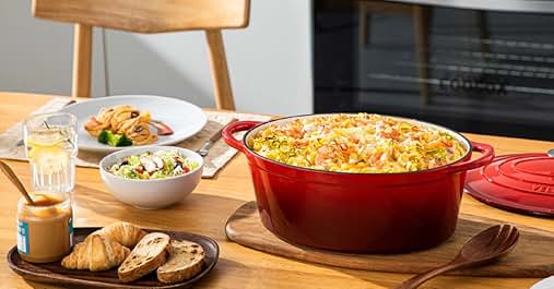 Large red oval casserole dish on wooden board, filled with cheesy pasta bake. Surrounding items include salad, bread, and drinks on a kitchen table.