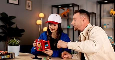 Dos personas jugando con un dispositivo educativo o de juego rojo sobre una mesa de madera, con iluminación ambiental y plantas de interior de fondo.