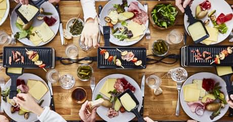 Vue aérienne d'une table avec plusieurs assiettes de fromage à raclette, de viandes, de légumes et de petites grillades. Mains visibles en train de chercher de la nourriture. Surface de table en bois avec différents plats et verres.