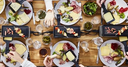Vue aérienne d'une table avec plusieurs assiettes de fromage à raclette, de viandes, de légumes et de petites grillades. Mains visibles en train de chercher de la nourriture. Surface de table en bois avec différents plats et verres.